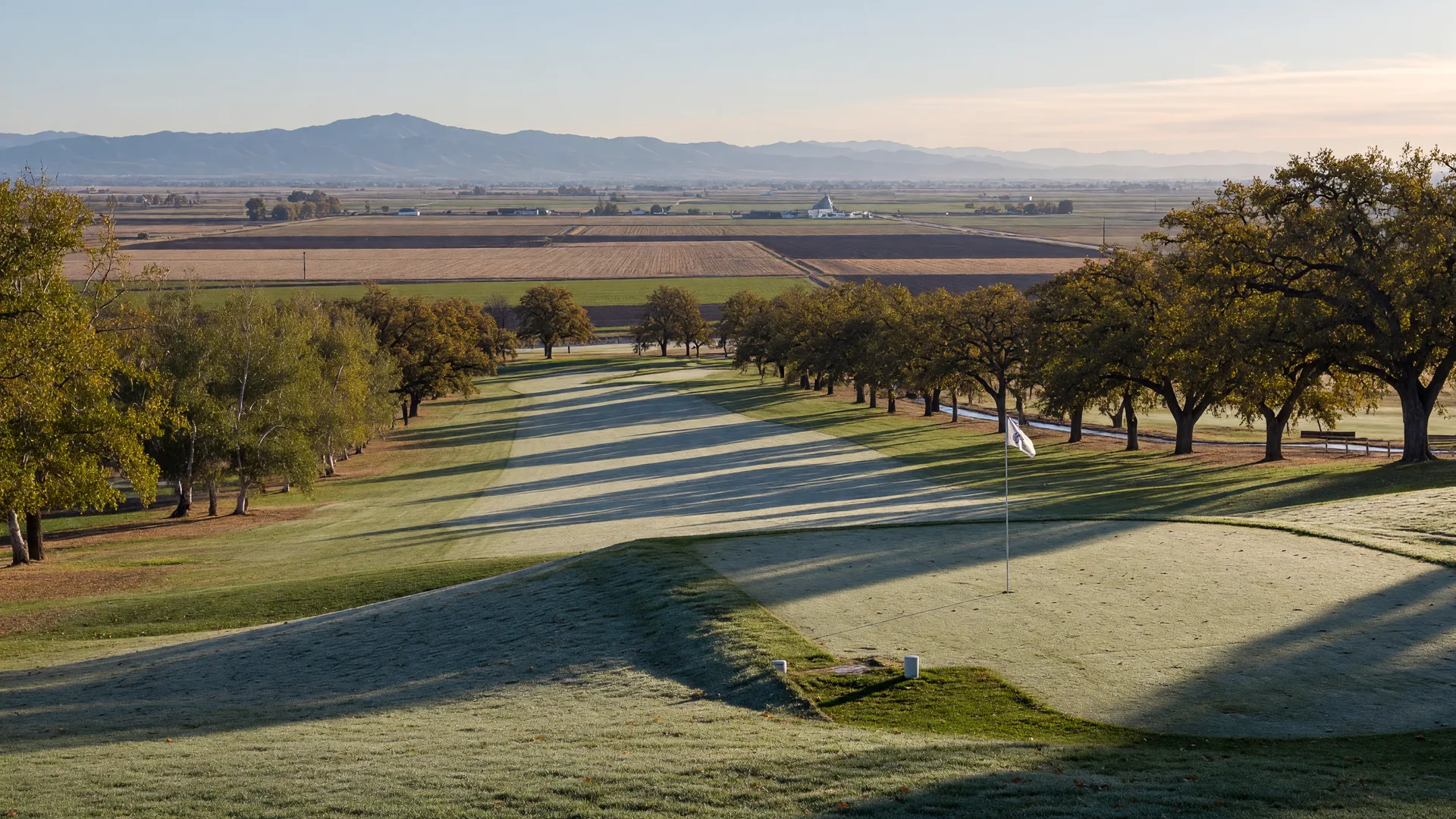 A classic Central Valley California parkland golf course on a crisp late-October morning, a long dew-covered fairway lined with valley oak and cottonwood trees, a green flag and white tee markers, low golden morning light casting long shadows, and the hazy Sierra Nevada foothills on the horizon under a clear pale-blue sky
