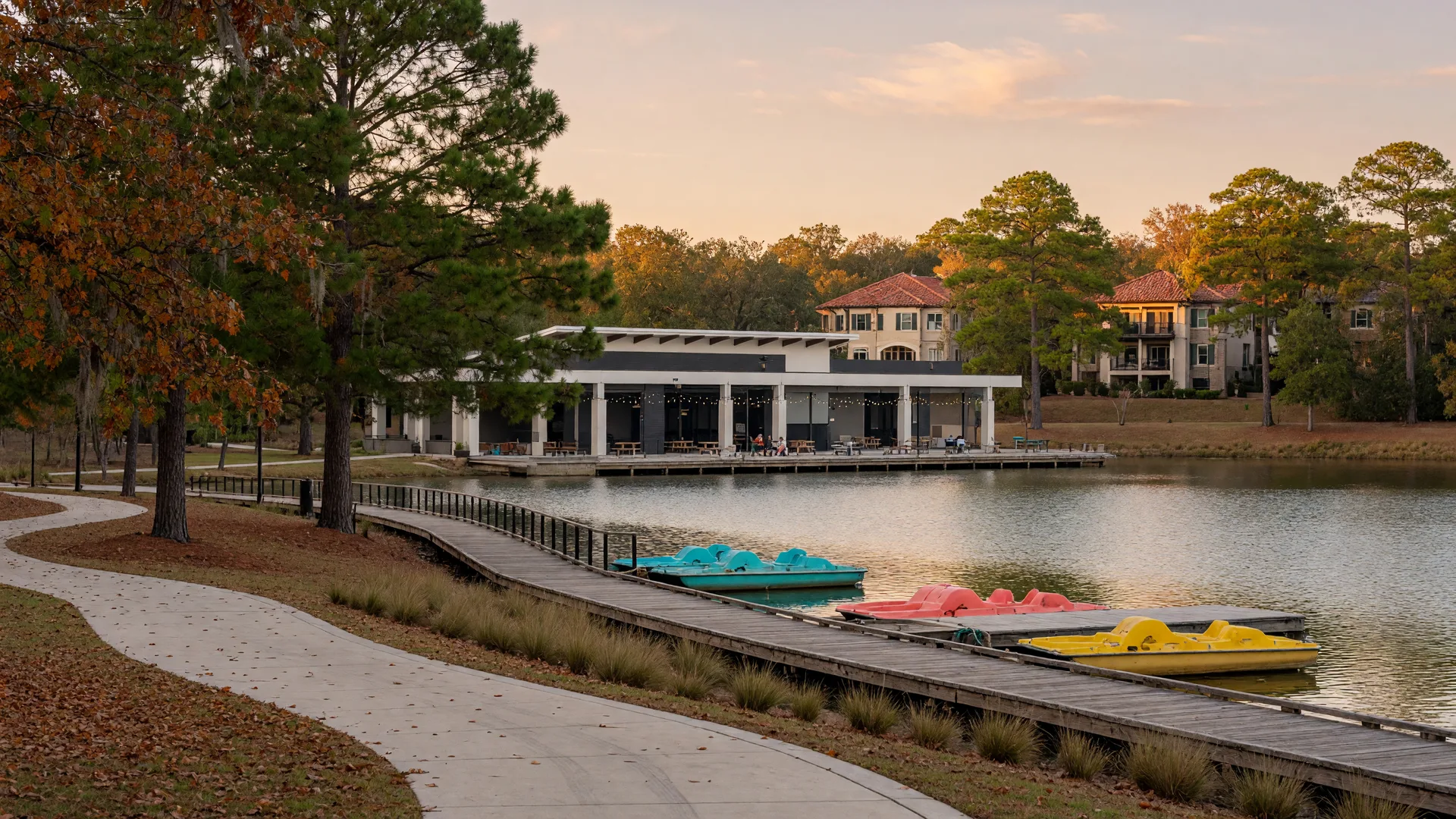 A boardwalk-lined lake with paddleboats at a Texas master-planned community amenity park in early autumn, warm light through loblolly pines