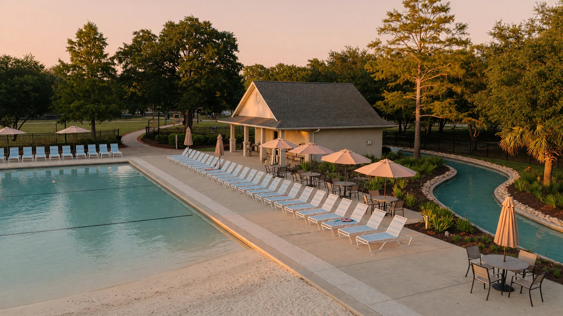 A wide editorial photograph of a community pool amenity area at golden hour, with a beach-entry pool, empty lounge chairs, and a low amenity building among trees