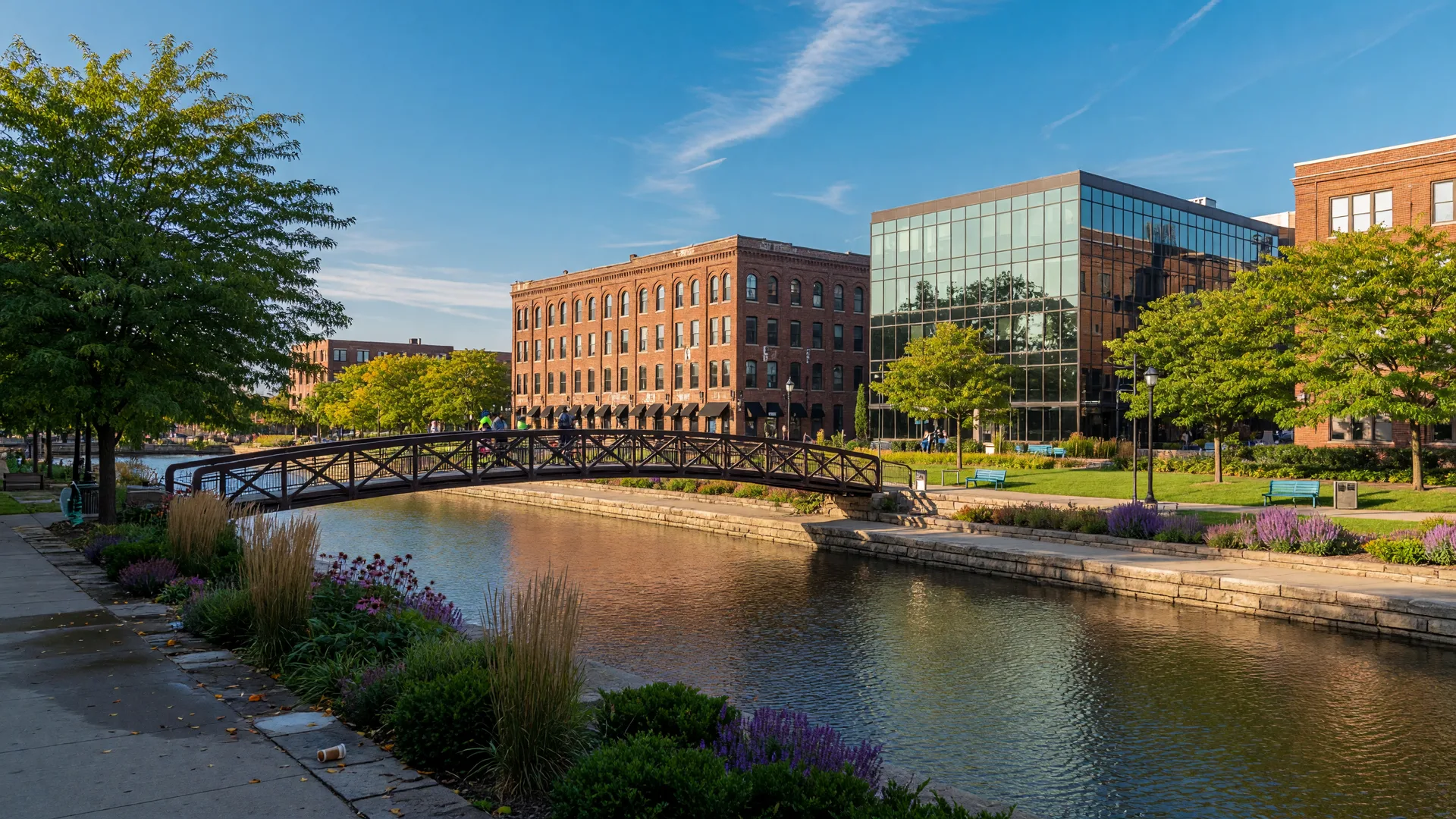 Downtown Flint, Michigan along the Flint River with restored brick buildings, a landscaped riverwalk, and a footbridge on a bright late-summer afternoon