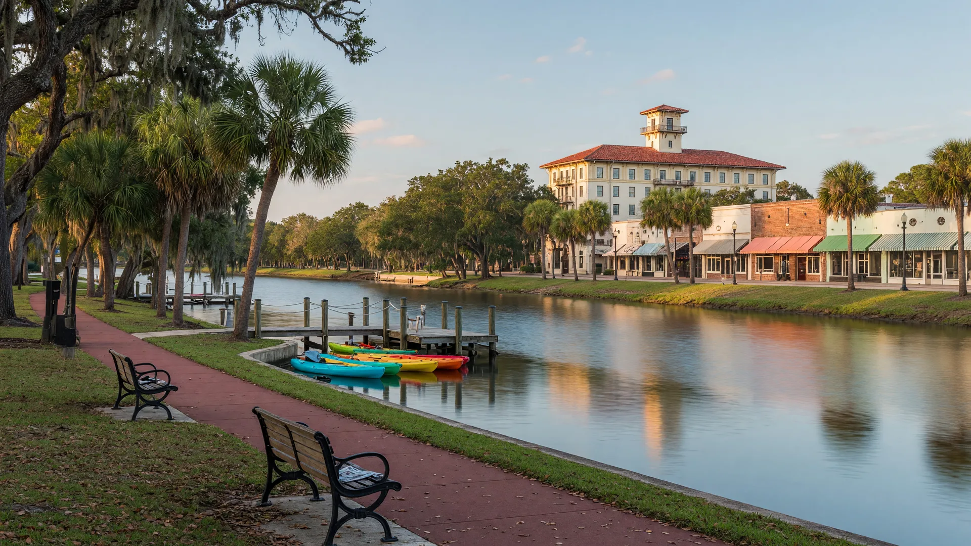 Downtown New Port Richey riverfront along the Cotee River on a clear late-summer morning, with a paved walking path, moored kayaks, moss-draped live oaks framing Sims Park, and the Hacienda Hotel rising behind the treeline