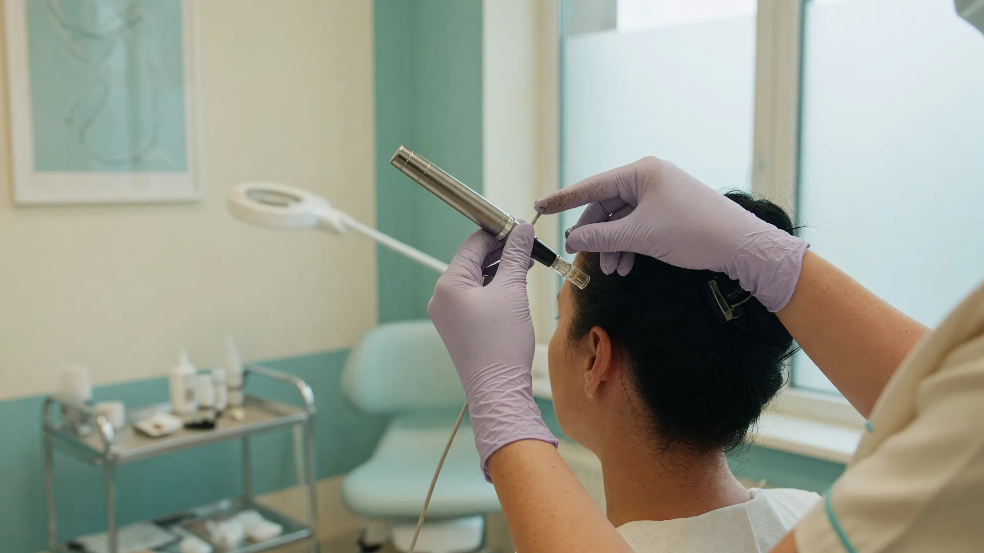 A clinician's gloved hands guiding a microneedling pen near the back of a patient's head in a bright treatment room