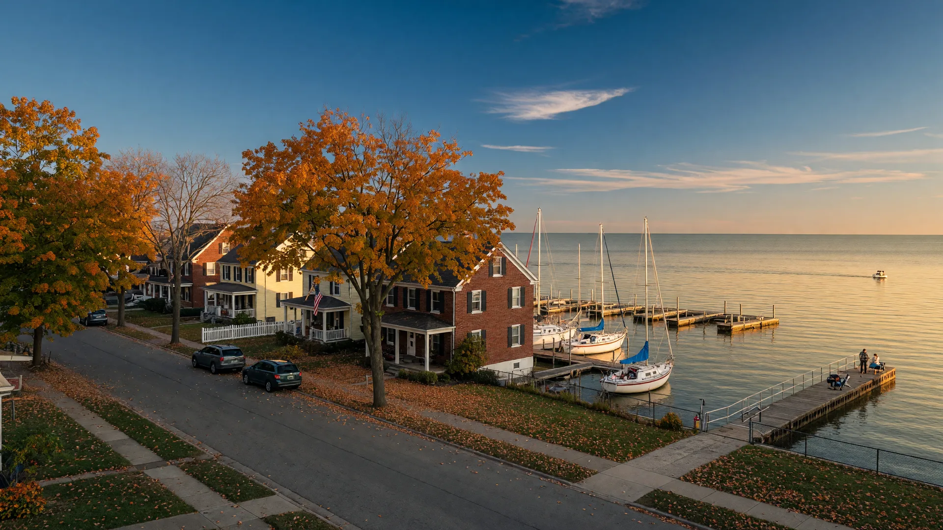 A Lake Erie shoreline street in Eastlake, Ohio in early autumn with brick and colonial homes under maples turning orange and gold, a small marina with sailboats and a fishing pier on the lake beyond in warm late-afternoon light