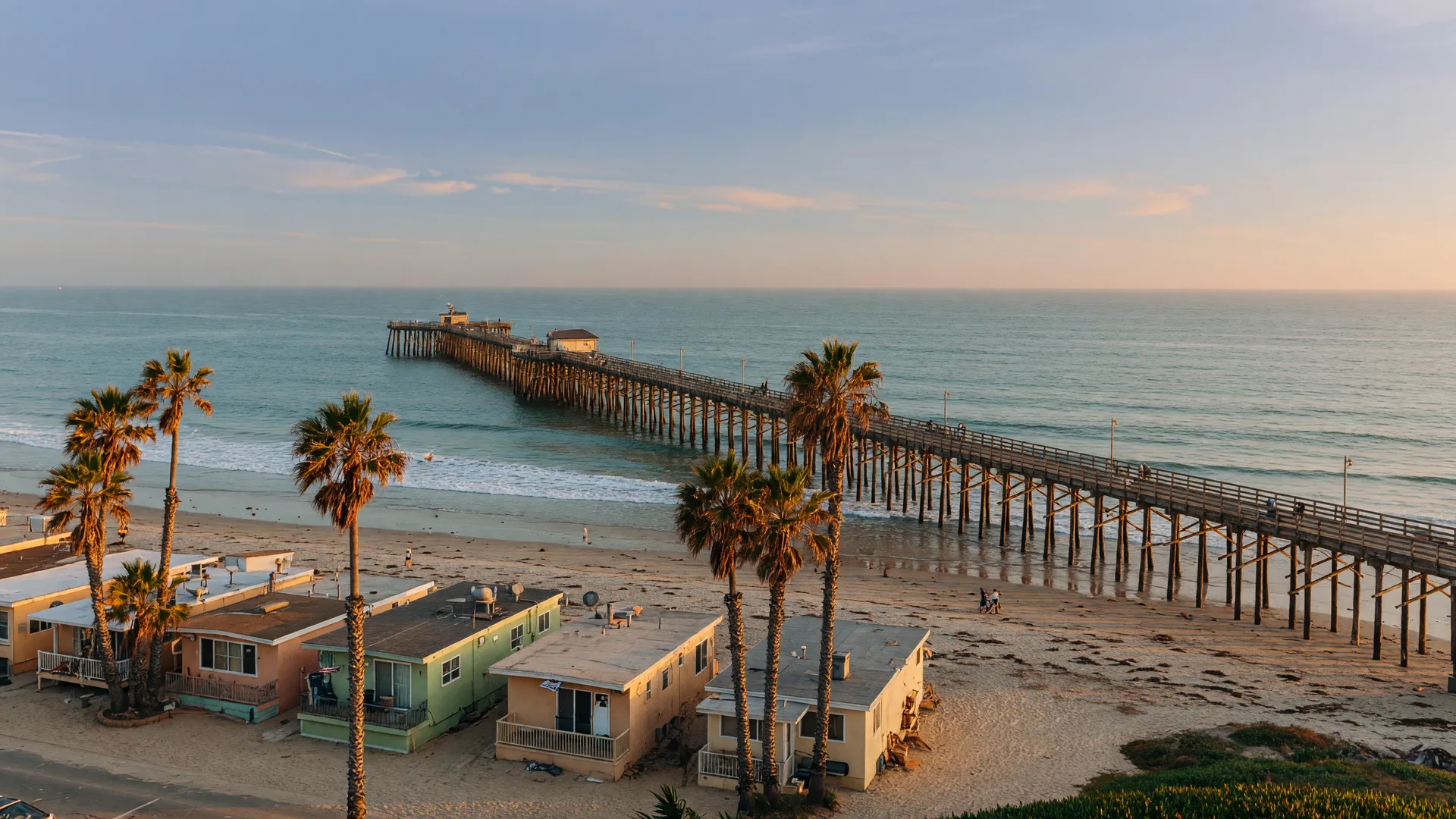 Golden hour view of the Imperial Beach Pier over the Pacific with low beach homes and palm trees along the shoreline in ZIP 91932