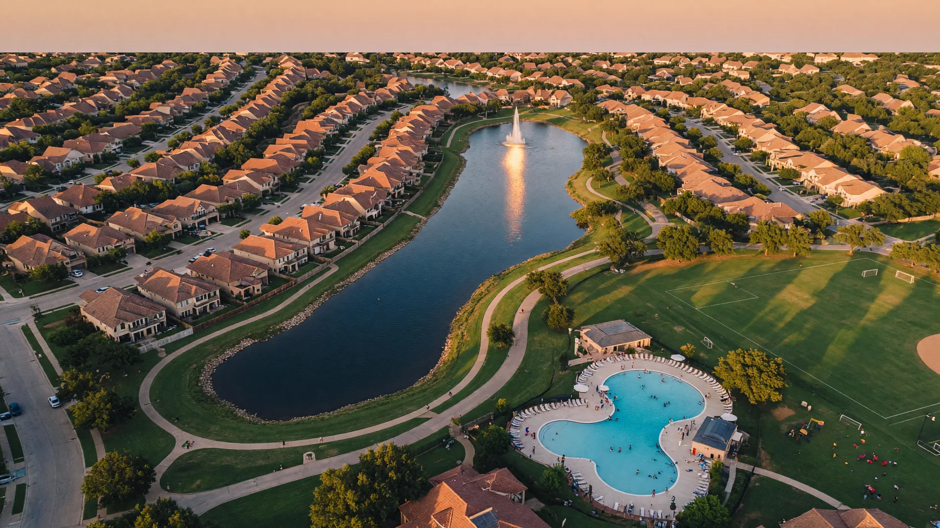 Aerial view of a Katy-area master-planned community at golden hour with a winding lake, trails, and a community pool