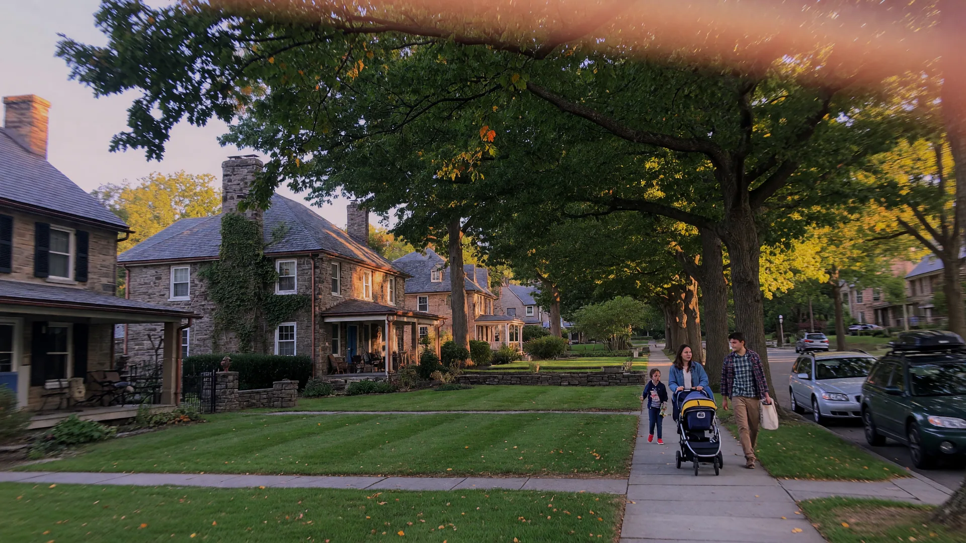 A tree-lined residential street in Rydal PA in mid-September with golden late-afternoon light across stone and brick homes under mature oaks with the first hints of autumn color