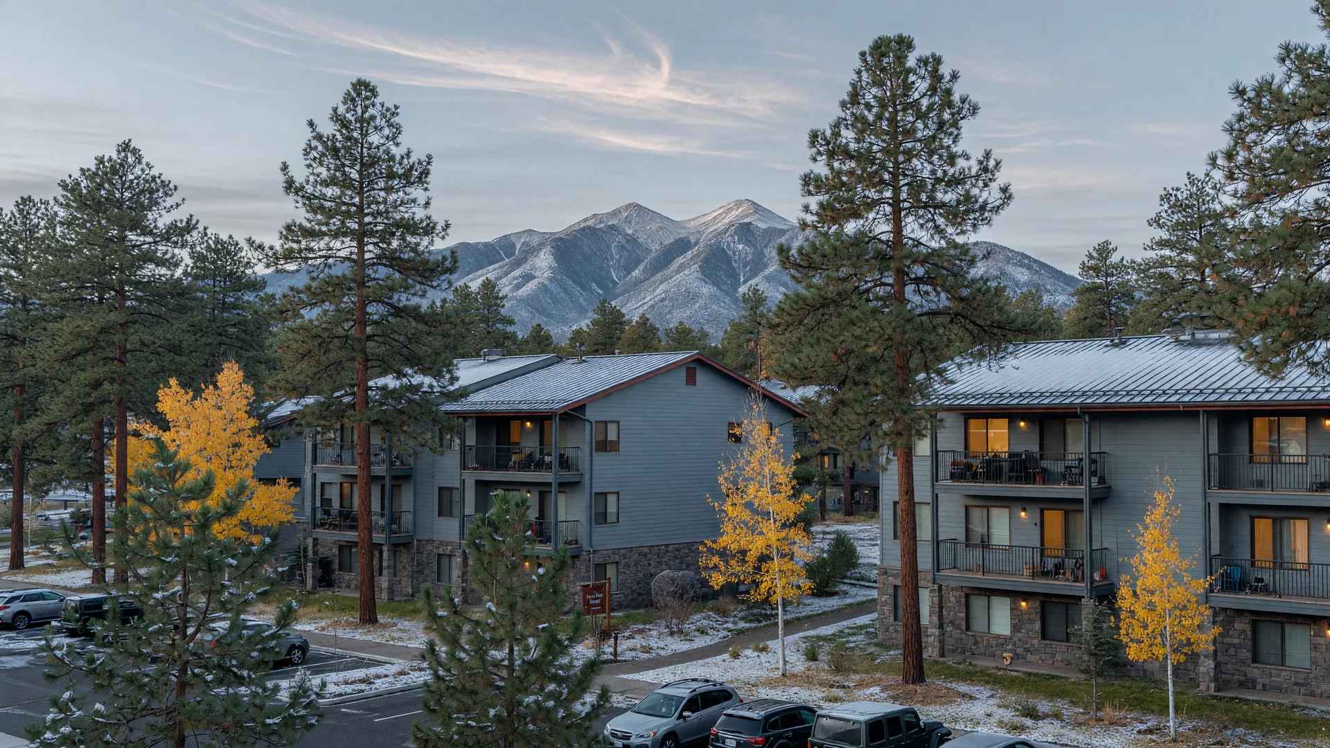 Modern mid-rise apartment buildings in Flagstaff with grey-blue siding among ponderosa pines and golden aspens, with the snow-dusted San Francisco Peaks in the distance on a late October morning