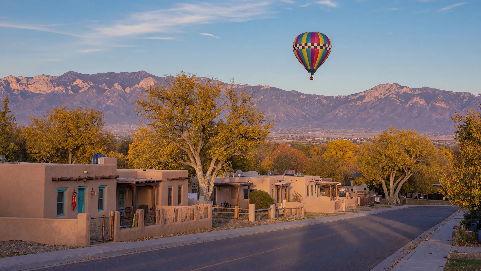 Golden autumn light on adobe pueblo homes with wooden vigas near Albuquerque, cottonwoods turning gold, the Sandia Mountains rising behind, and a hot air balloon drifting over the Rio Grande valley