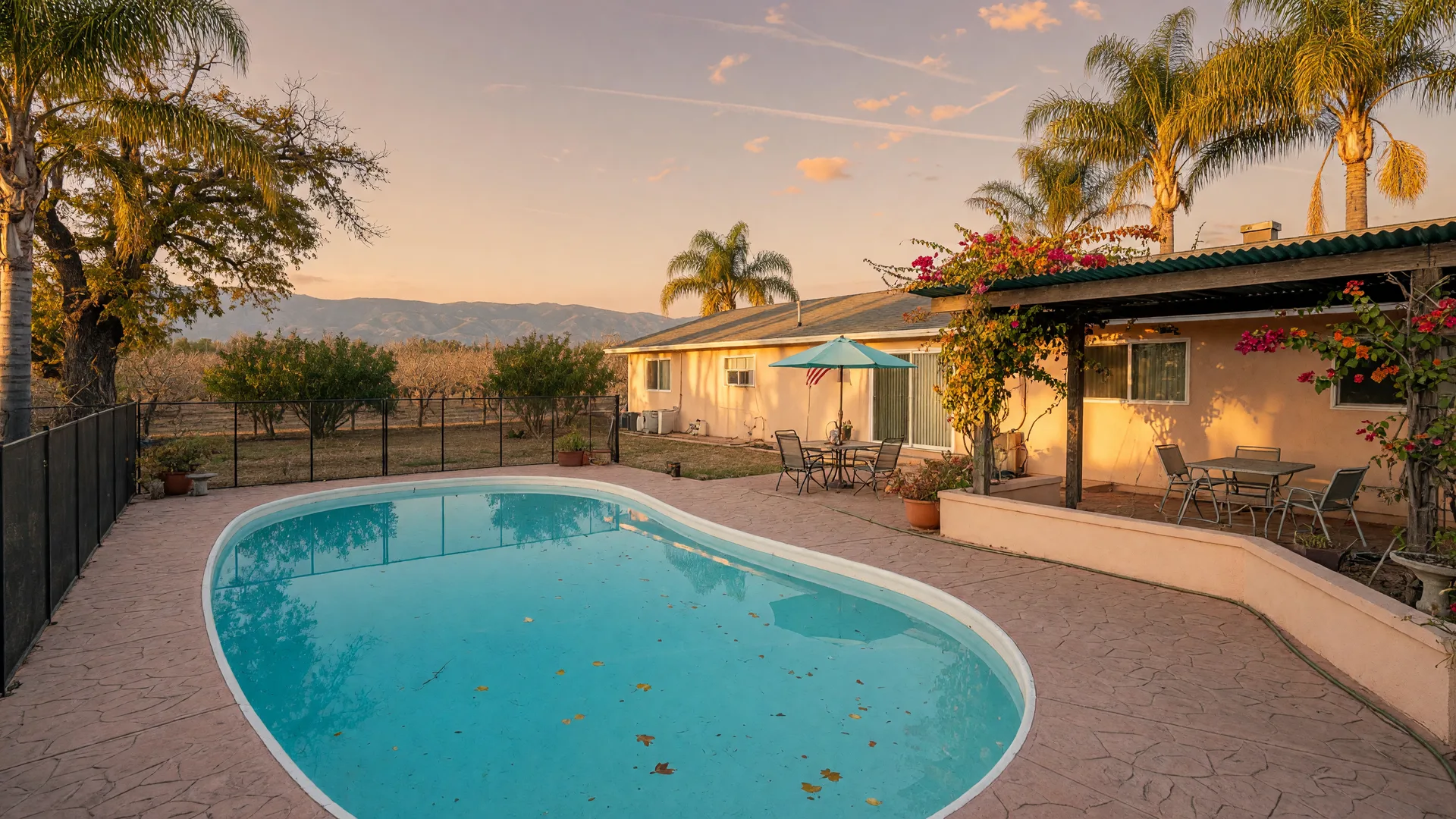 A backyard in-ground swimming pool at a single-story ranch home in Merced County on a warm October afternoon, clear turquoise water, a stamped-concrete patio under a covered pergola, palm trees and a valley oak, and the hazy Sierra Nevada foothills on the horizon