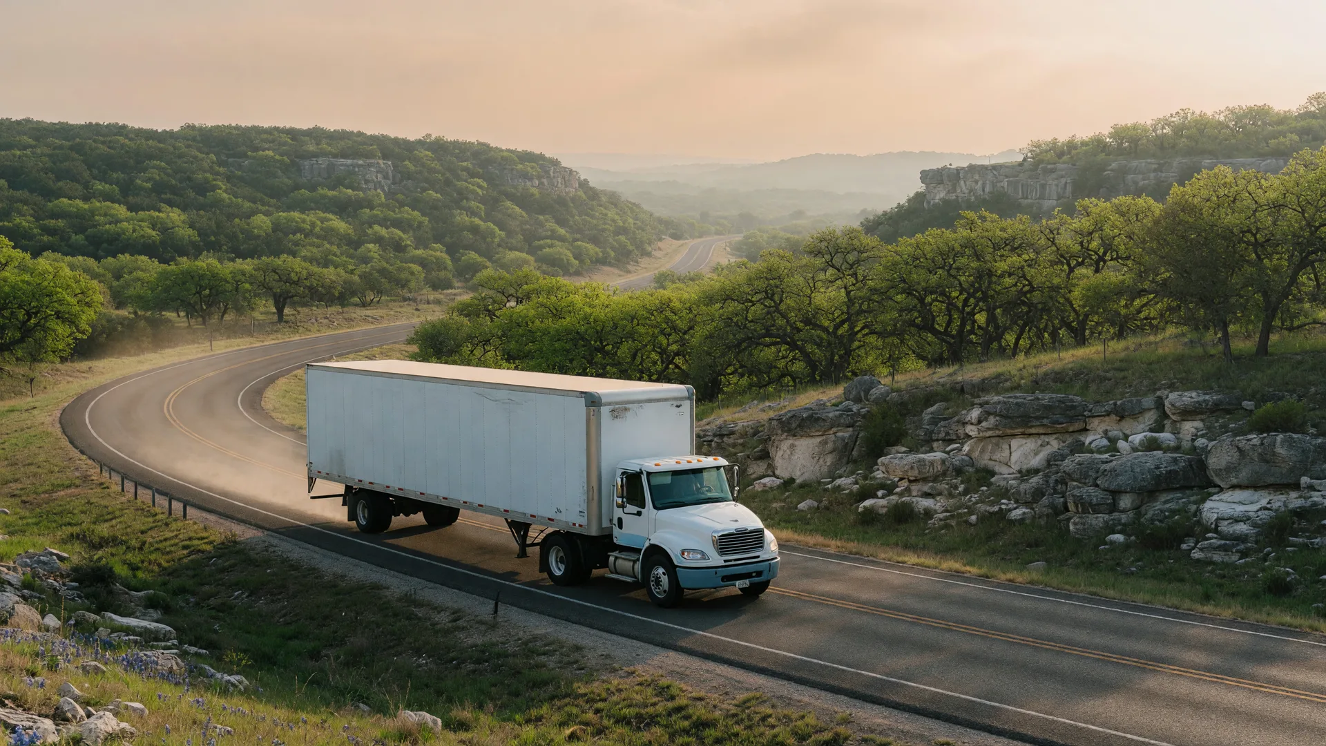A moving van on a two-lane Texas Hill Country highway through rolling green hills in morning light