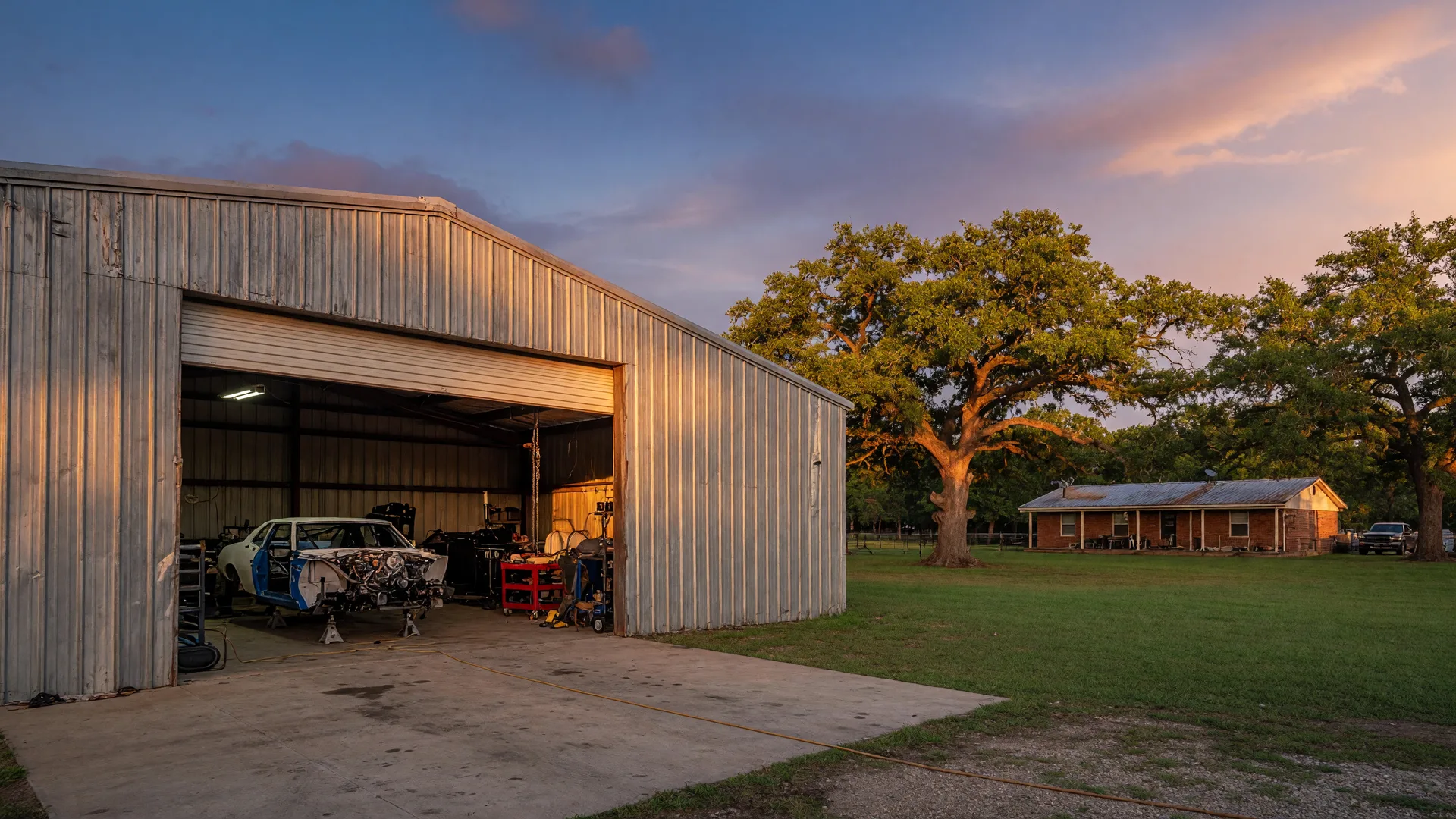 A large metal workshop on a rural Northeast Texas property at golden hour with a race car project inside the shop through an open roll-up door, surrounded by pasture and oak trees