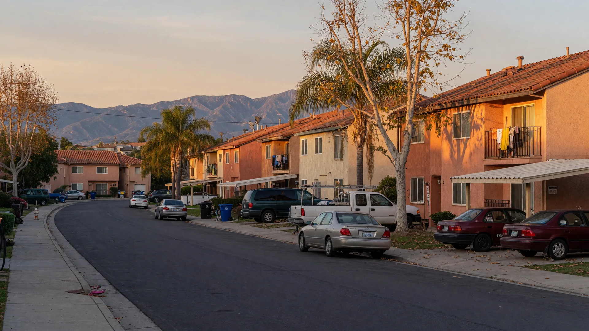 A quiet residential street in Valencia, Santa Clarita lined with apartments and townhomes, palm trees, and parked cars at golden hour, with mountains on the horizon