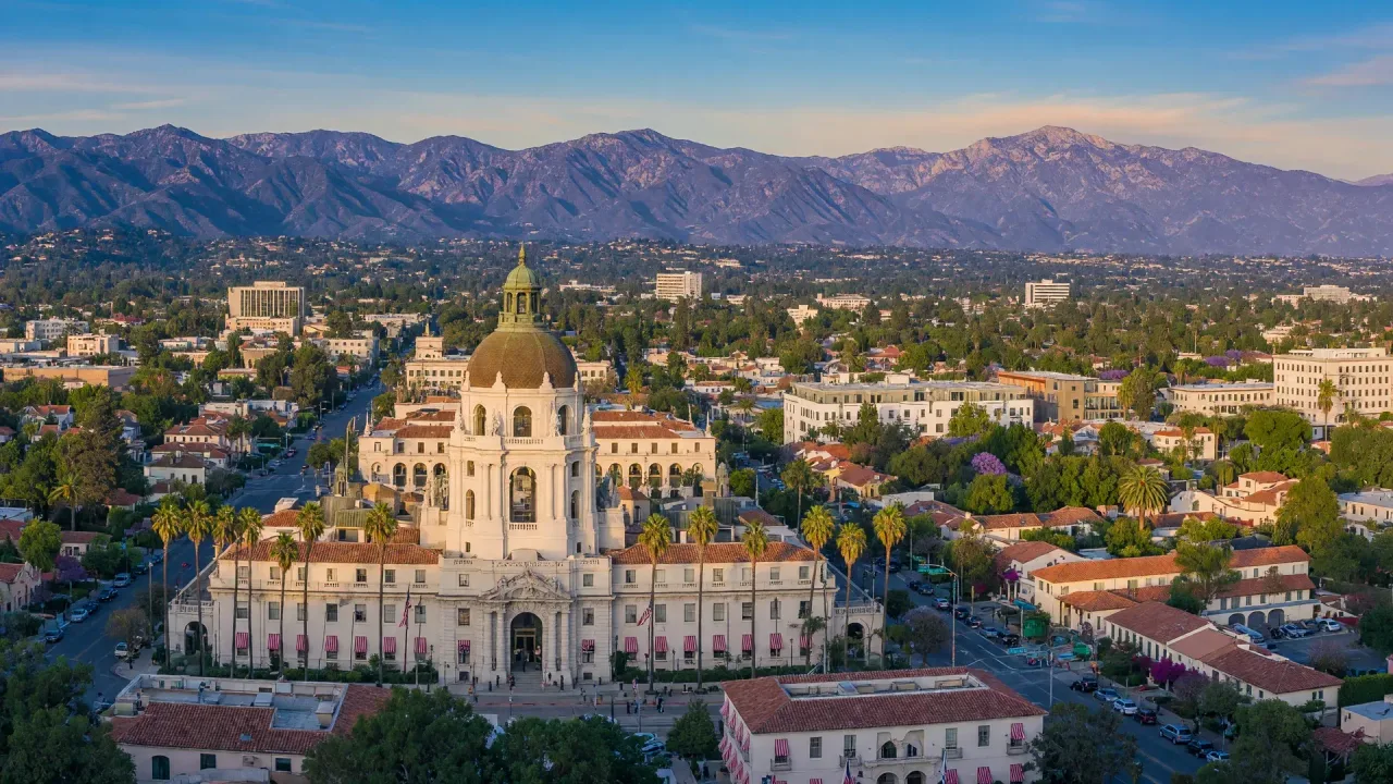 A wide postcard-style panorama of Pasadena, the skyline and the San Gabriel Mountains beyond under a warm sky