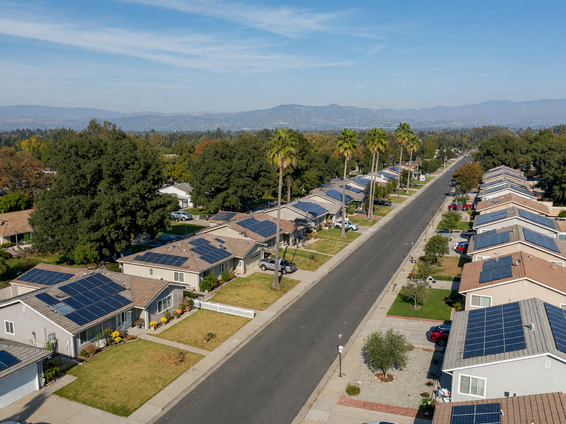 An elevated view of a Merced County residential street in early fall with ranch and tract homes, several rooftops carrying solar panel arrays, neat lawns, oak and palm trees, and hazy Sierra Nevada foothills on the horizon