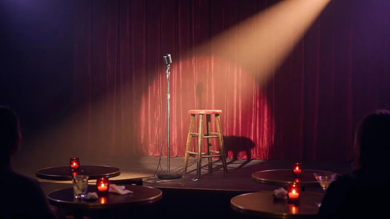 An intimate comedy club stage set for a stand-up show, a vintage microphone and wooden stool under a tight spotlight with a red curtain backdrop and dark tables in the foreground