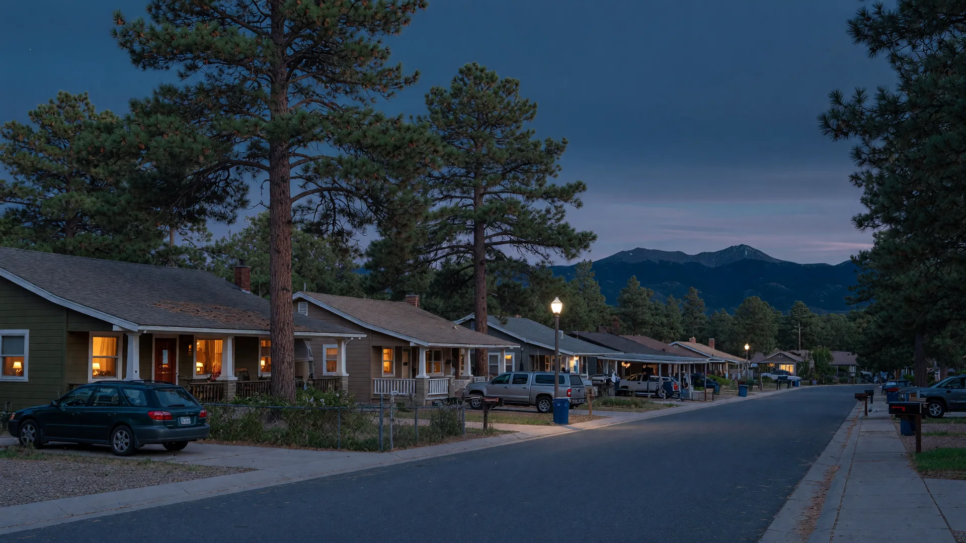A Flagstaff neighborhood street at blue hour in October, warm lights glowing in home windows beneath ponderosa pines and the San Francisco Peaks