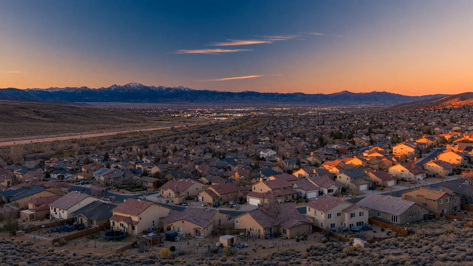 A golden-hour view of Sparks, Nevada, with rooftops in the foreground, faint city lights beginning to glow, and the Sierra Nevada range on the horizon.