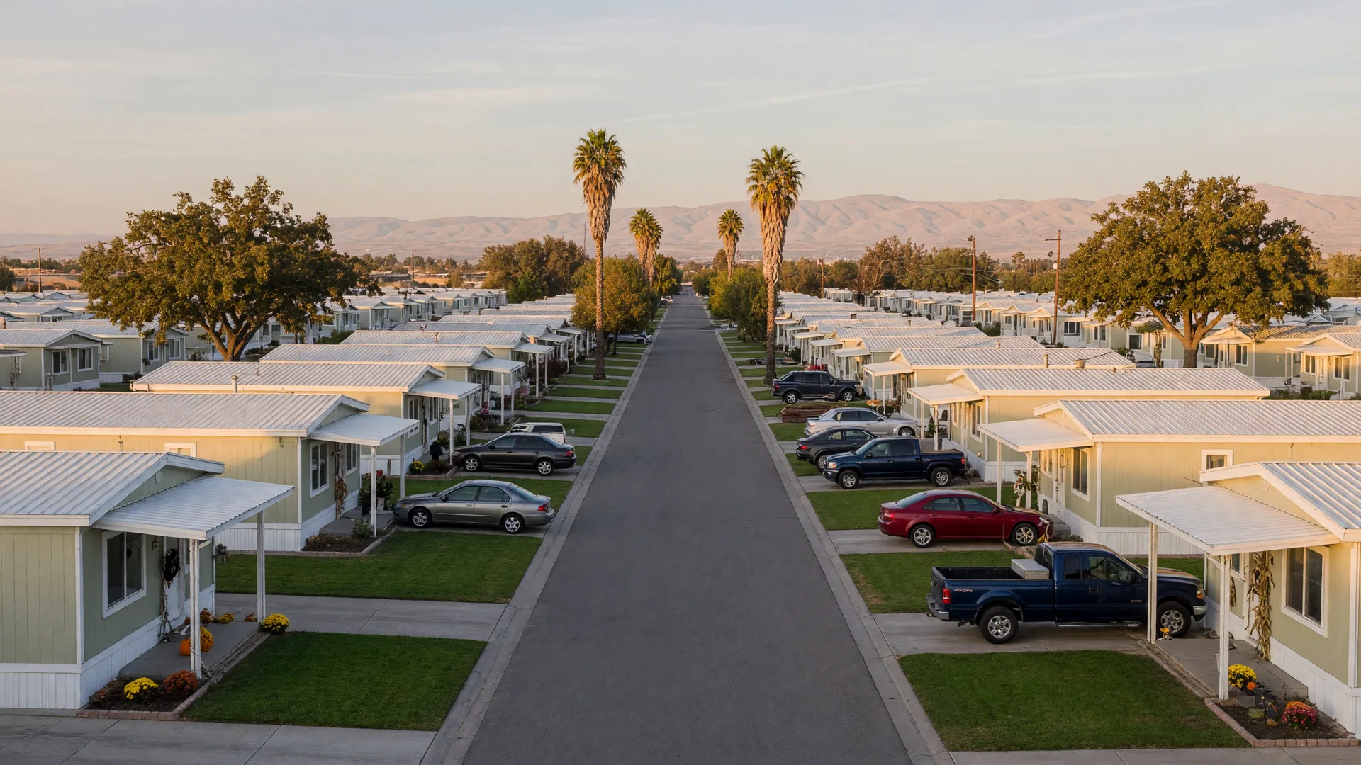 A tidy manufactured home community in the Central Valley on a warm mid-October late afternoon, long rows of vinyl-skirted homes with covered porches and neat lawns, oak and palm trees, and the hazy Sierra Nevada foothills on the horizon