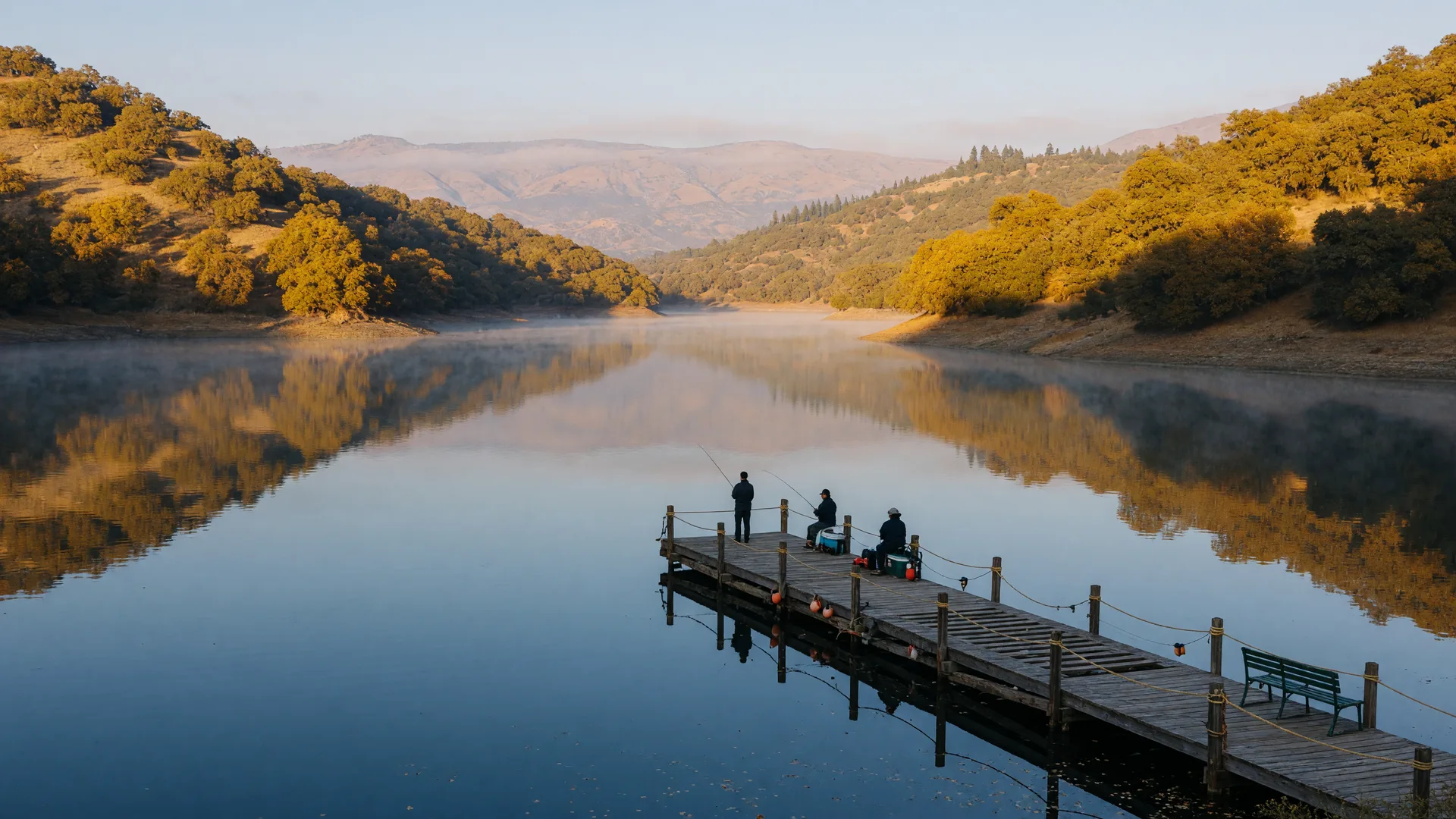 Lake McSwain in the Merced County foothills on a crisp October morning, calm water reflecting golden oak-covered hillsides, a wooden fishing dock, and the hazy Sierra Nevada foothills on the horizon