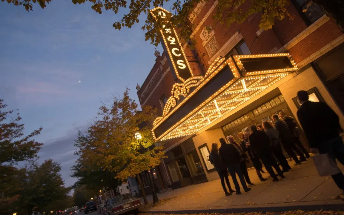The historic Keswick Theatre in Glenside at dusk with its marquee glowing warm against a blue-hour sky and a crowd gathering beneath the canopy on a tree-lined street