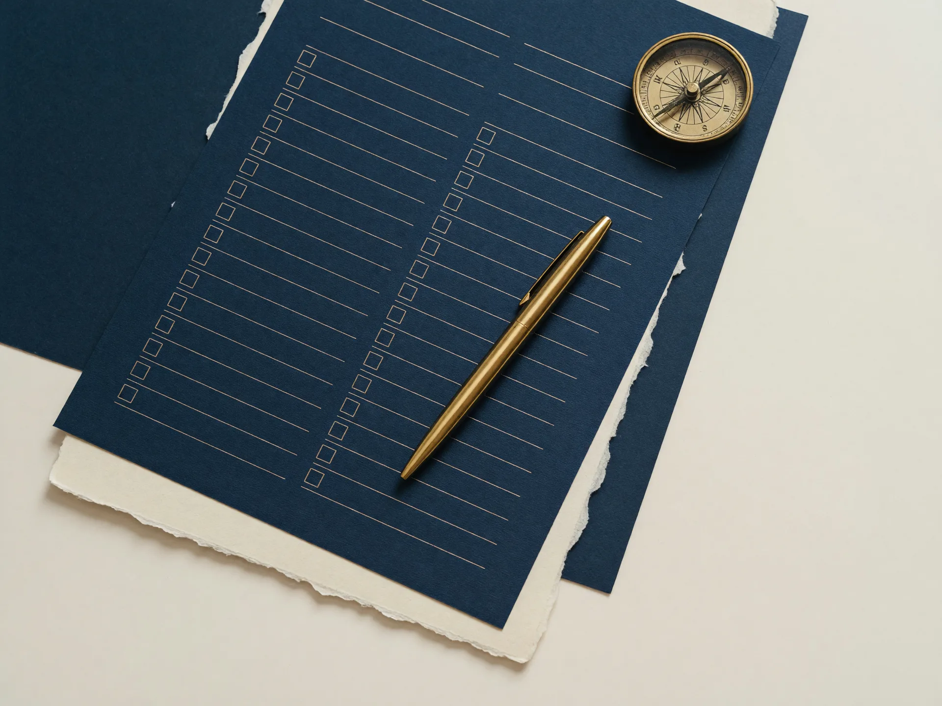 A navy checklist worksheet with gold ruled lines and blank check boxes, a gold pen, and a small brass compass