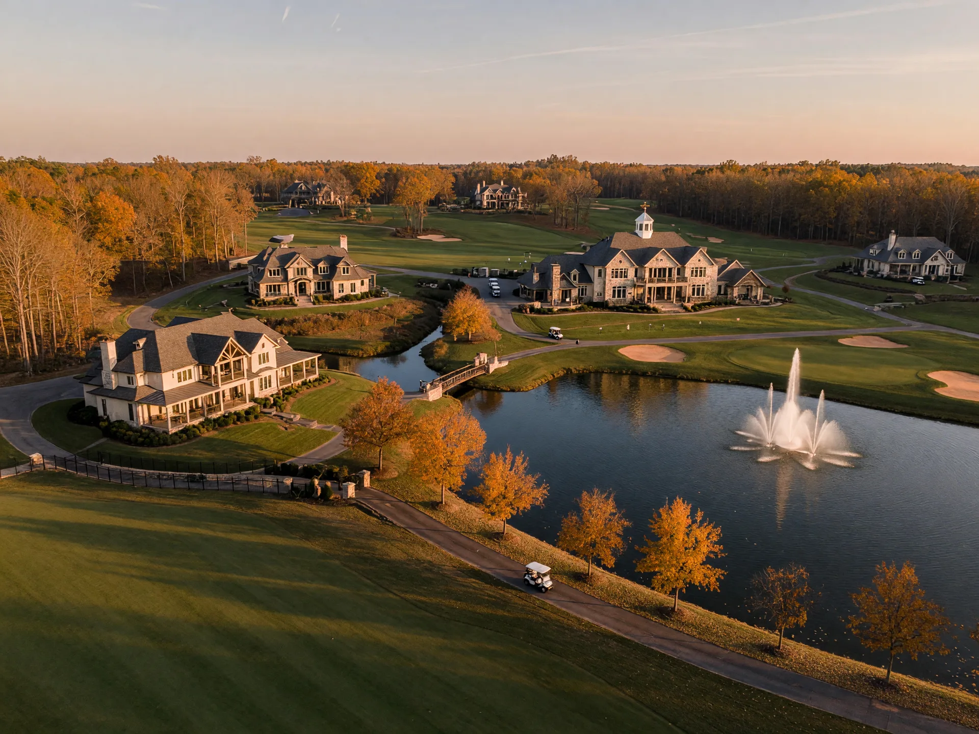 Aerial drone view of a master-planned luxury golf community in Northeast Georgia in late autumn with sculpted fairways, a fountain lake, estate homes, and a clubhouse in warm afternoon light