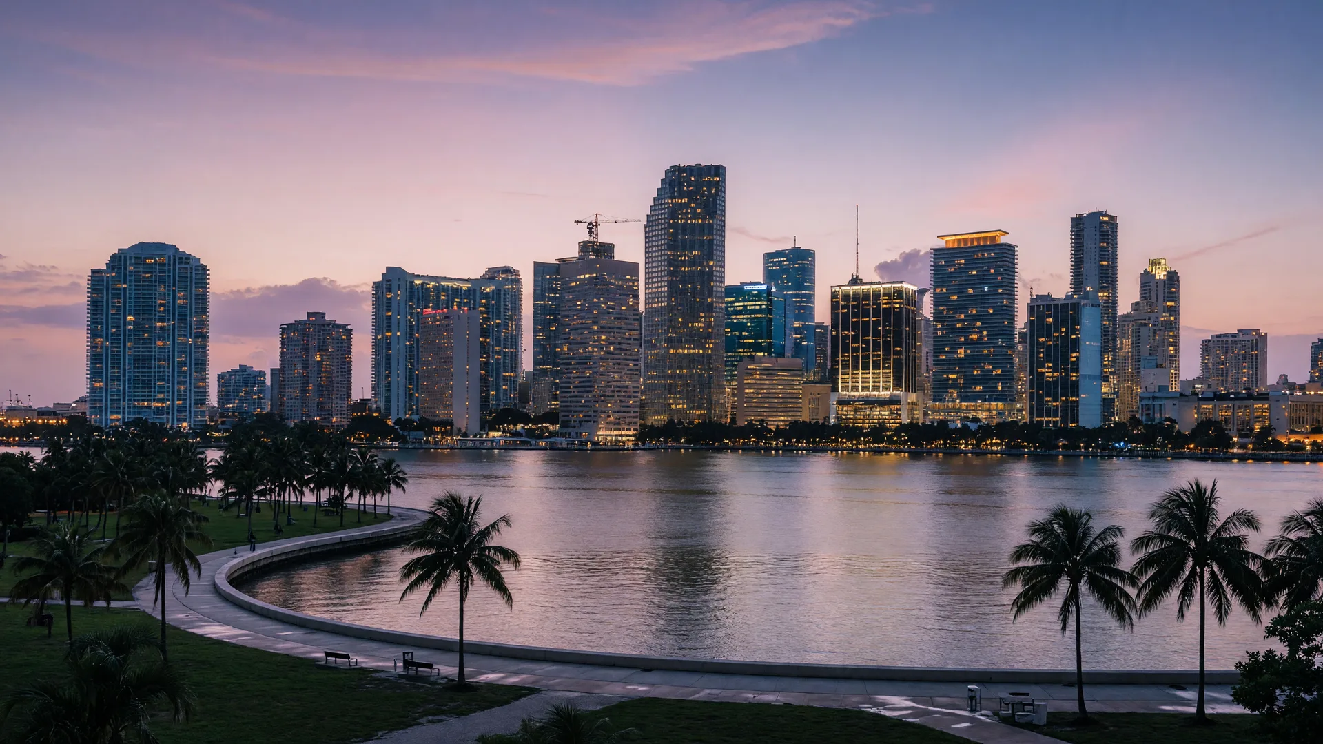 The downtown Miami skyline across Biscayne Bay at dusk, with palms silhouetted along the bayfront