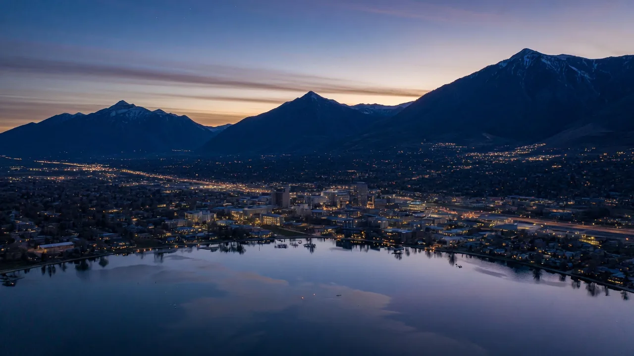 Utah Valley at dusk with the lights of Provo and Orem spreading across the valley floor, Utah Lake in the foreground, and Mount Timpanogos silhouetted on the east.