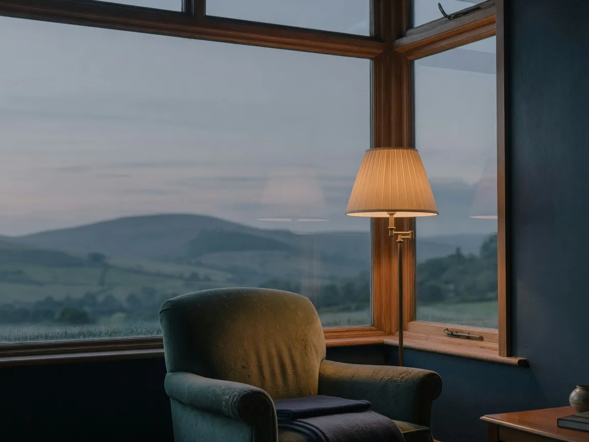 A calm view through a large window at dusk over quiet rolling hills, a warm armchair in the foreground