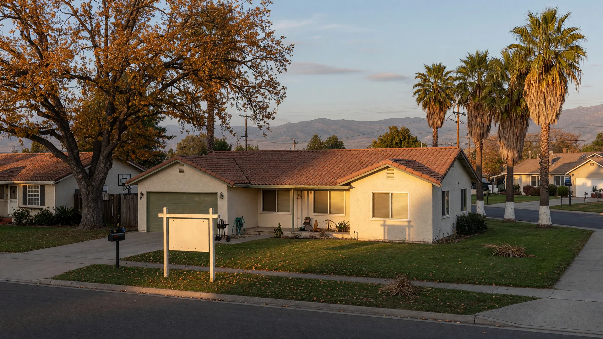 A well-kept single-story cream stucco ranch home in Merced County at golden late-October afternoon light, with a plain wooden real estate sign standing in the front yard, a neat green lawn, valley oak and palm trees along the quiet street, and the hazy Sierra Nevada foothills on the horizon