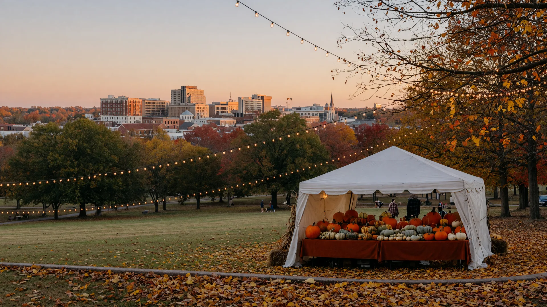 Festival tents, pumpkins, and warm fairy lights in a Southern park at golden hour with a distant city skyline and fallen autumn leaves