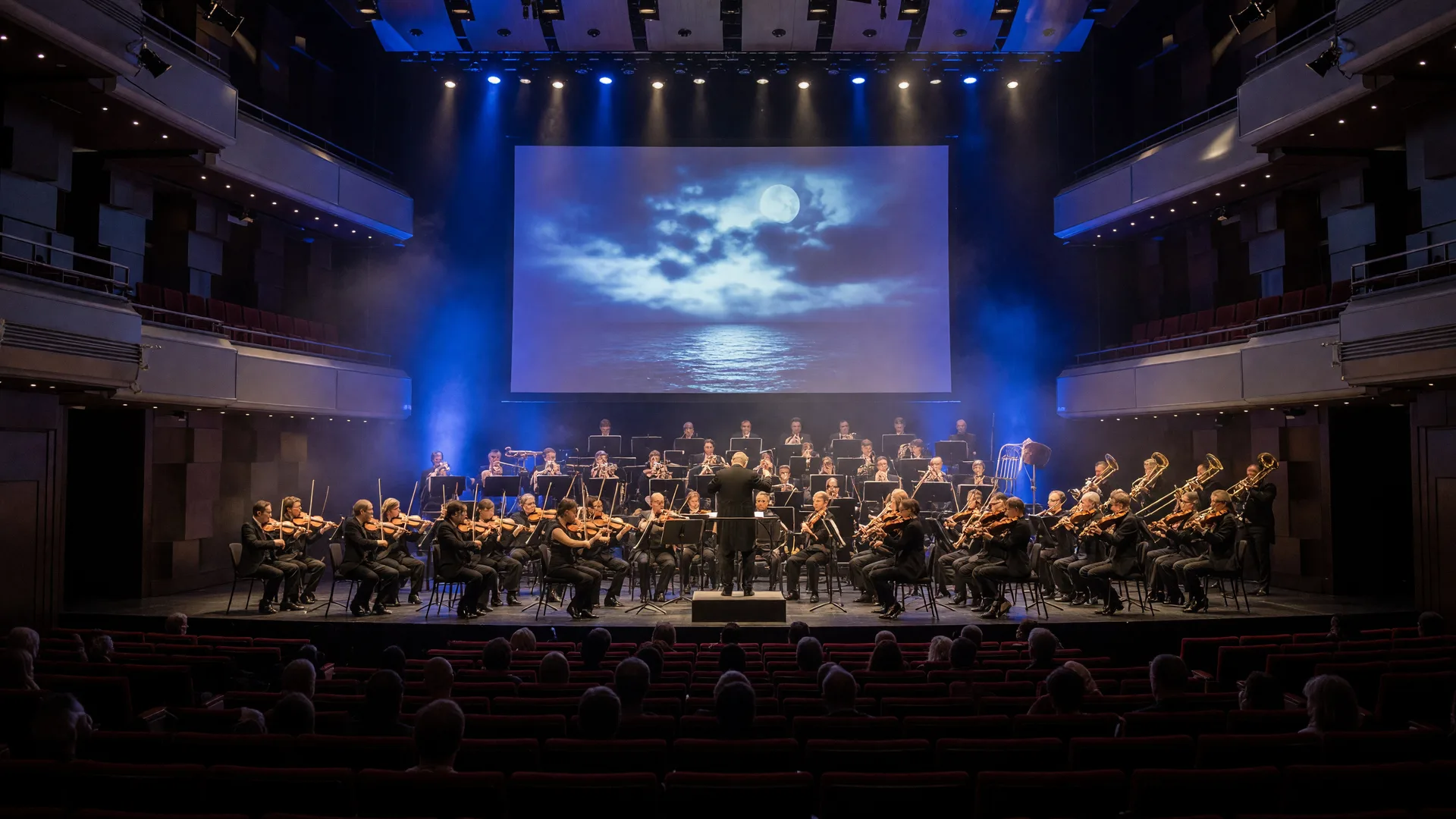A symphony orchestra performing a film score concert in a modern concert hall with a warmly lit stage in deep blue and gold and a large glowing screen behind the orchestra