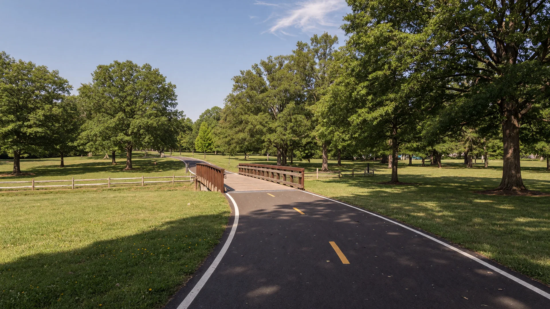 A flat paved multi-use recreation trail lined with mature trees and open parkland on a bright afternoon in Arlington, Virginia