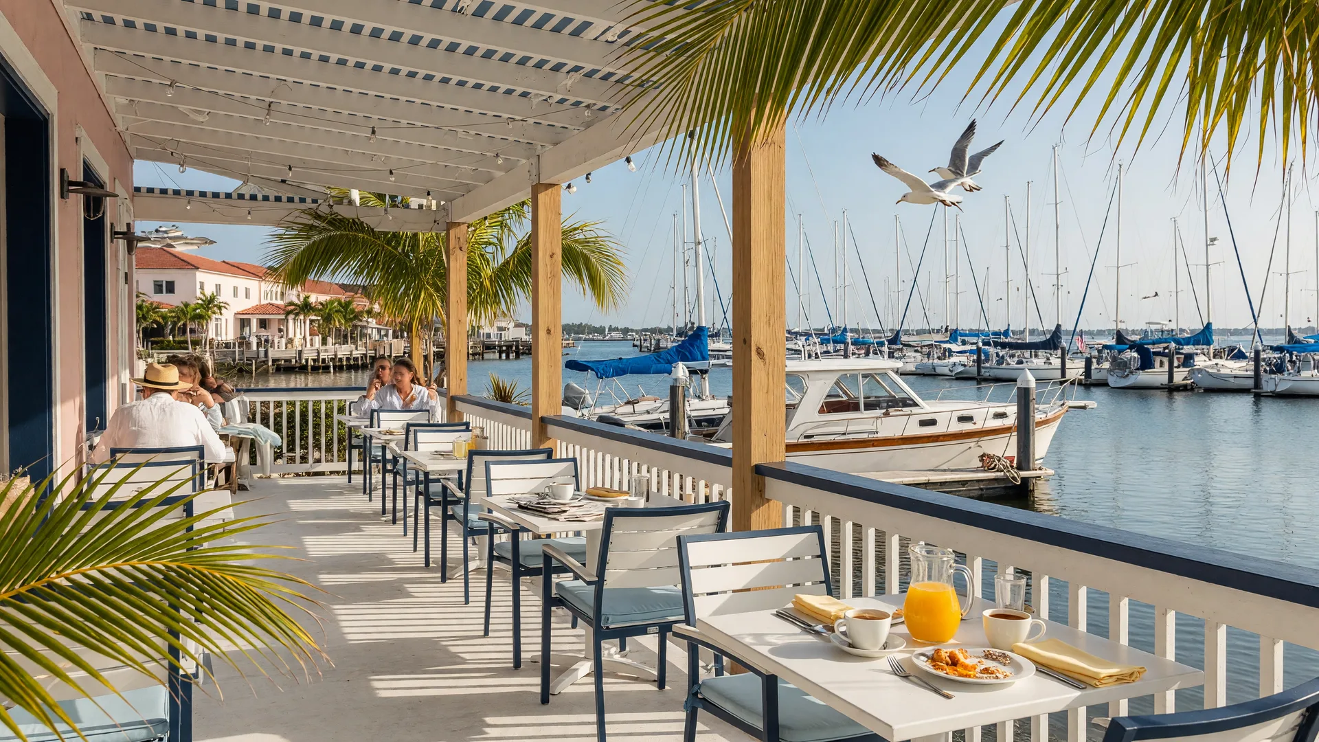 Bright waterfront breakfast cafe in Old Naples, Florida with a covered patio facing a calm marina, white tables, palm fronds, and warm morning light