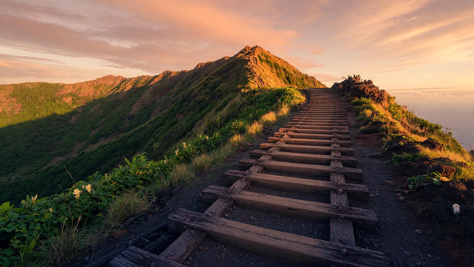 A golden-hour view of the Koko Crater Railway Trail on Oahu: a steep stairway of old railroad ties ascending a volcanic ridge with lush green slopes and trade-wind clouds