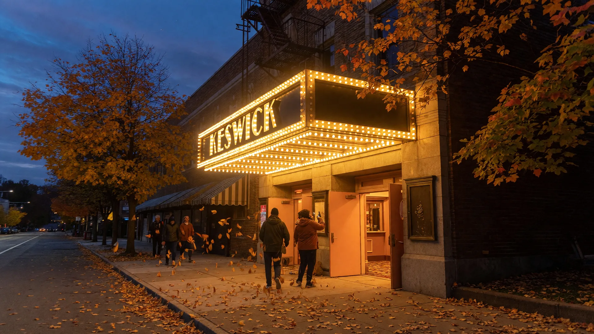 The Keswick Theatre in Glenside PA at dusk in early October, its glowing golden marquee lit against a deep blue-hour sky, warm light spilling from the lobby doors, autumn leaves drifting across the sidewalk, and distant figures walking toward the entrance