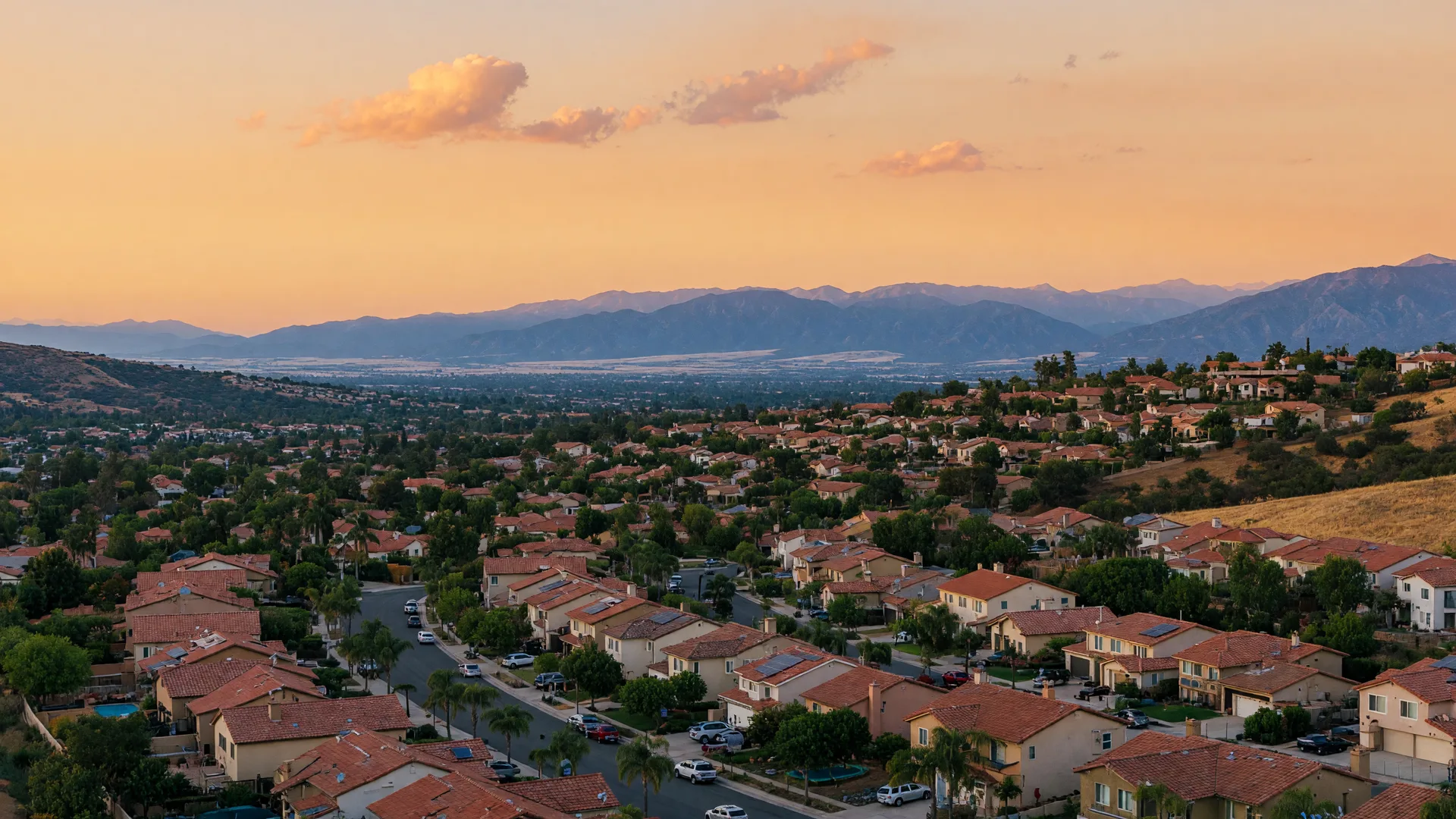 A golden-hour view across the Santa Clarita Valley: red-tile-roofed hillside neighborhoods, tree-lined streets, and the hazy San Gabriel mountains beyond