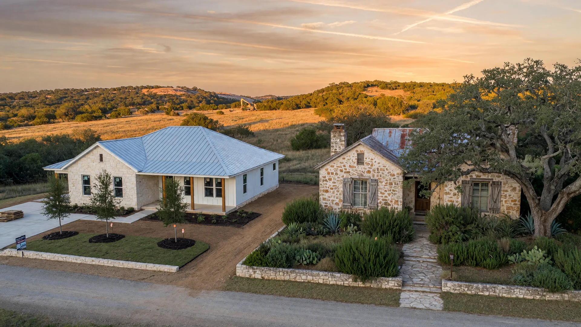 Side-by-side contrast of a brand-new limestone-and-metal Hill Country home with young oaks and an established limestone-and-cedar home with mature live oaks at golden hour near Boerne, Texas