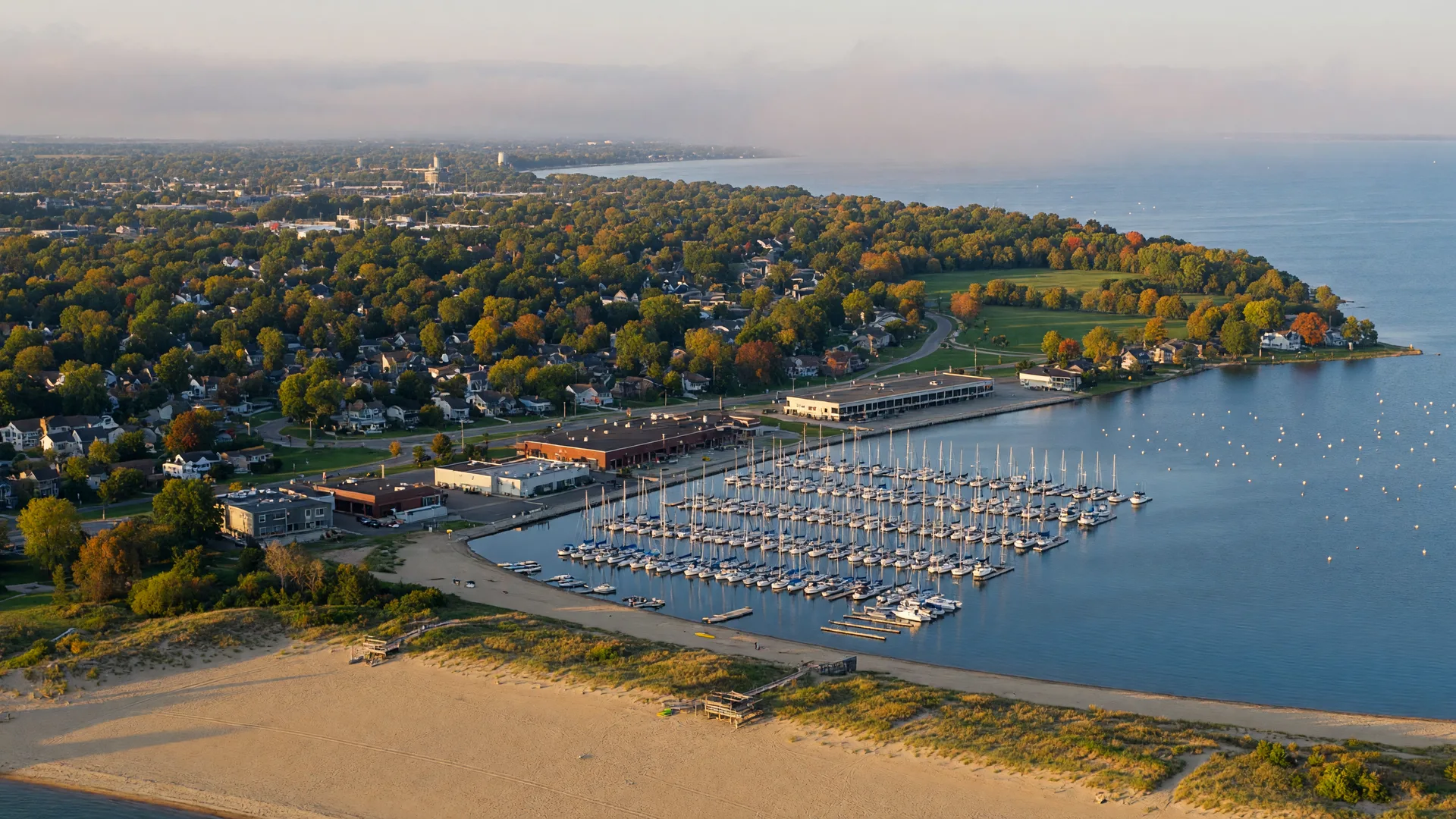 A wide aerial view of Mentor, Ohio on the southern shore of Lake Erie in early autumn, with a long sandy beach and marina in the foreground, sailboat masts on calm blue water, and tree-lined suburban neighborhoods turning gold and orange in warm late-afternoon light