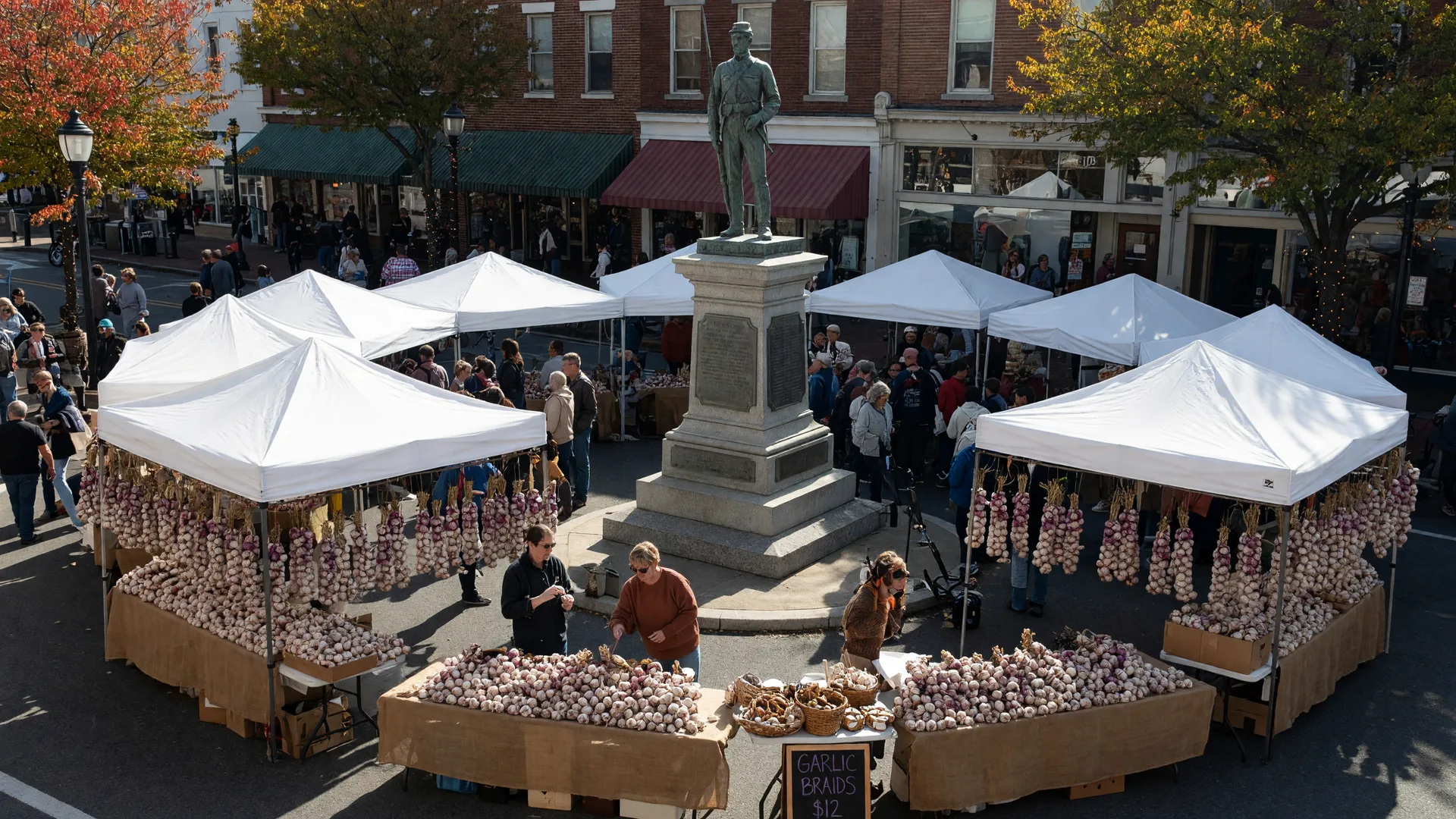 Centre Square in downtown Easton on Garlic Fest weekend, white vendor tents ringing the Soldiers and Sailors monument, stall tables piled with garlic bulbs and braided garlic strings, brick storefronts with awnings and red-and-gold autumn trees behind
