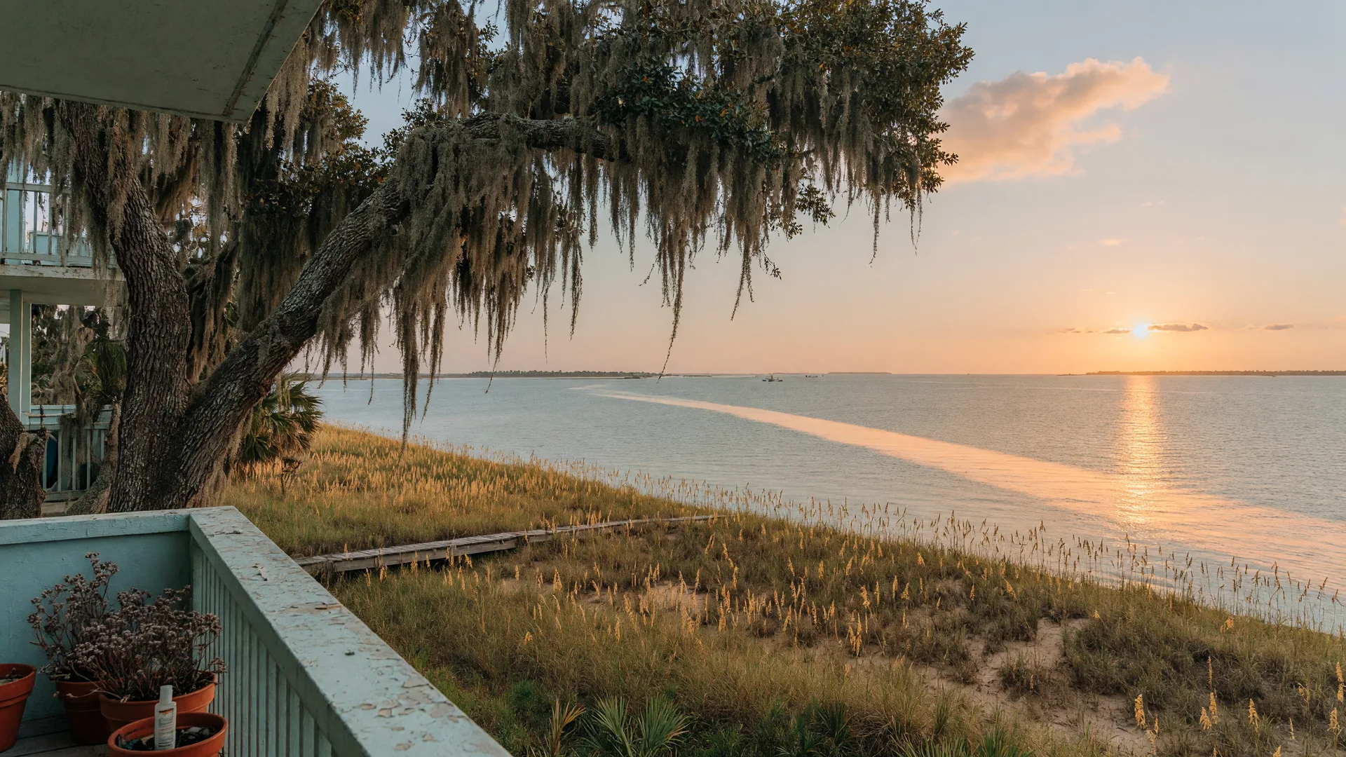 A Gulf-front condominium balcony overlooking the Mississippi Sound on a golden October afternoon, with sea oats and sand dunes in the foreground and a live oak draped in Spanish moss framing the scene