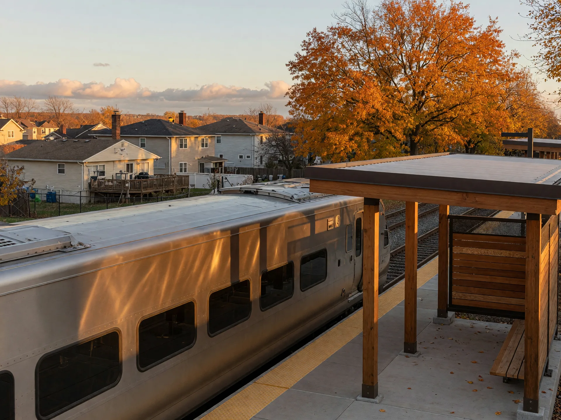 Modern electric commuter train at a new suburban rail station platform in Northwest Indiana on a clear fall afternoon, with a wood-accented station shelter and autumn trees behind it