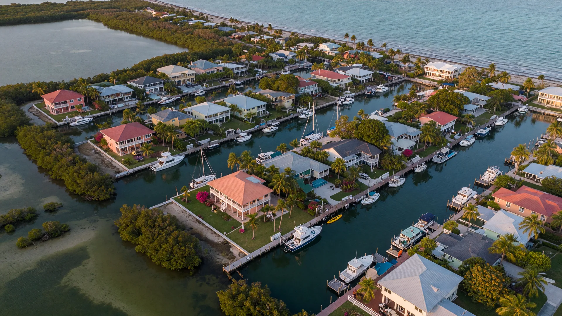 High aerial view of a Key Largo waterfront neighborhood at golden hour, a grid of deep-water canals lined with luxury elevated homes, private docks and boats, tropical lots, the Atlantic shore on one side and mangrove flats on the other