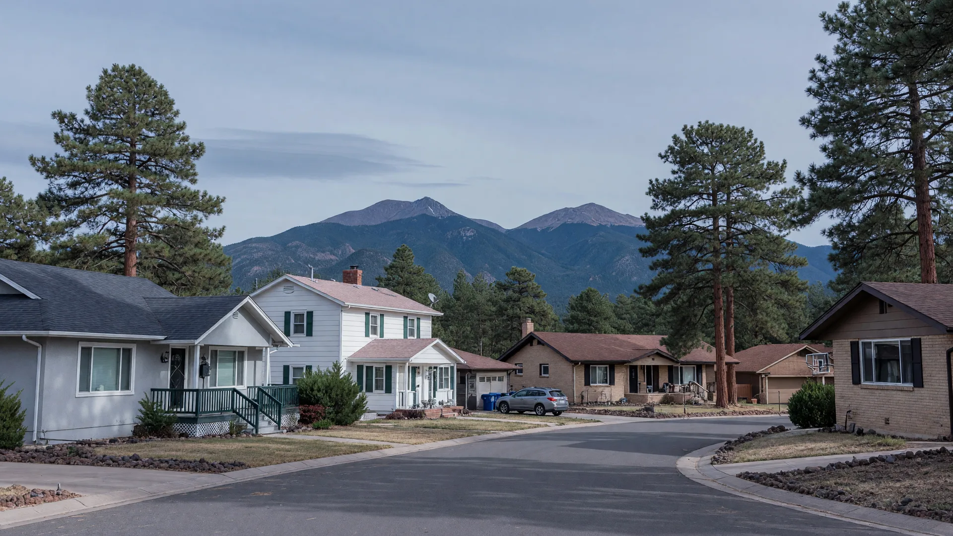 Flagstaff residential homes with pitched roofs among tall ponderosa pines under a cool blue-grey October sky with the San Francisco Peaks in the distance