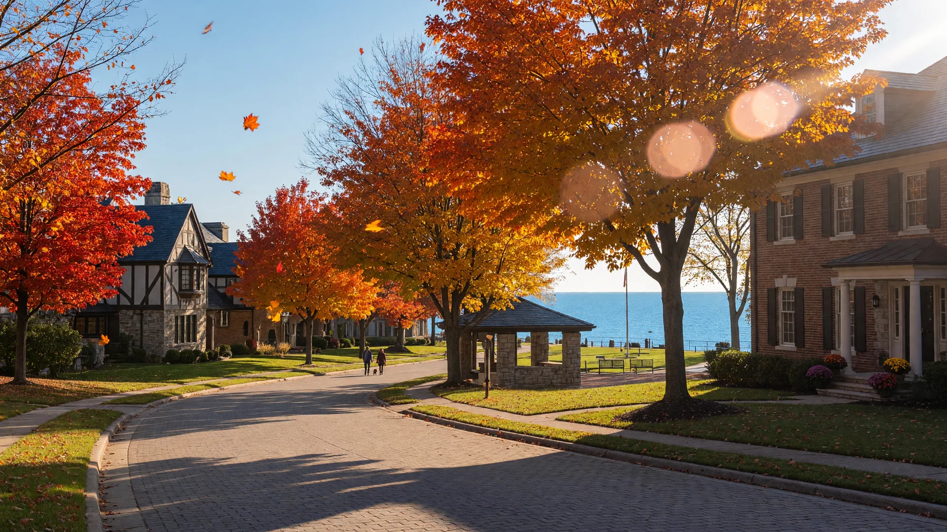 Tree-lined street of brick and stone luxury homes in Westlake, Ohio on a crisp October morning, sugar maples at peak autumn color, with a glimpse of Lake Erie shoreline park in the distance