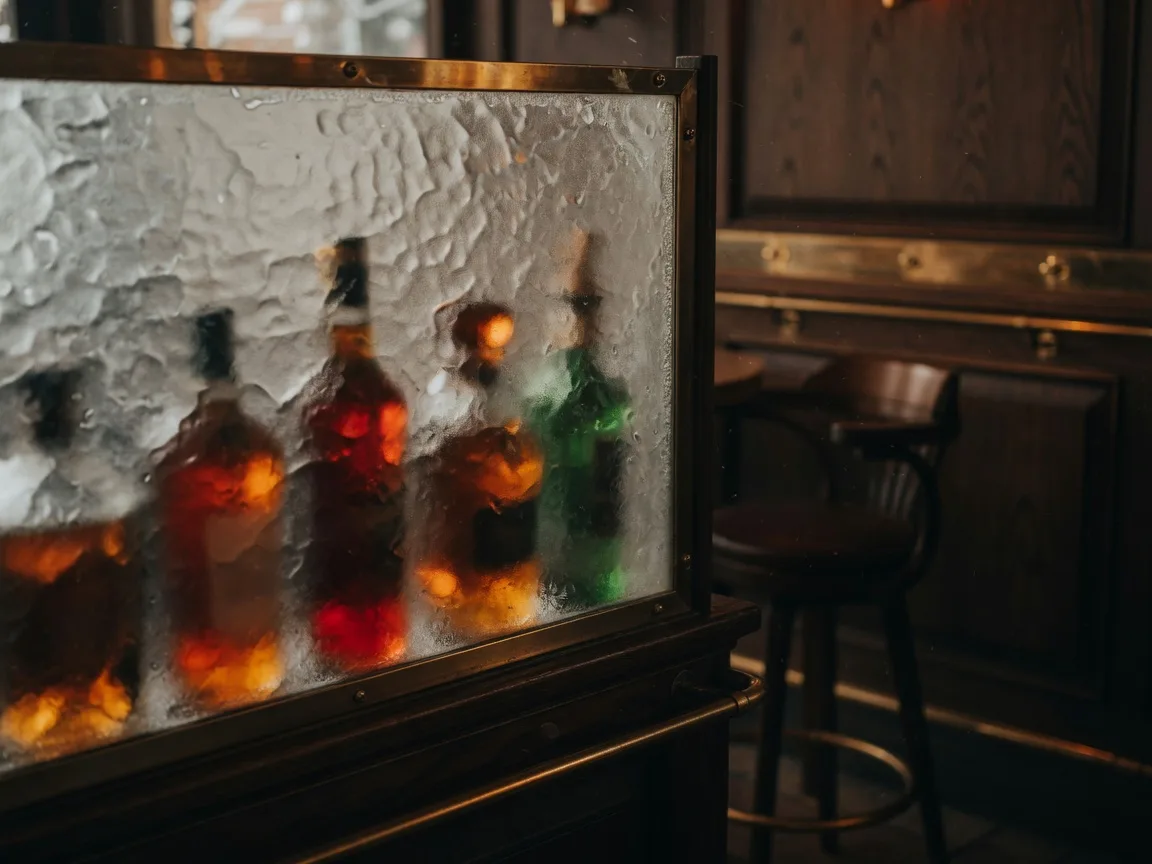 A frosted-glass partition glowing with warm amber light inside a dim upscale restaurant bar, bottles silhouetted behind it
