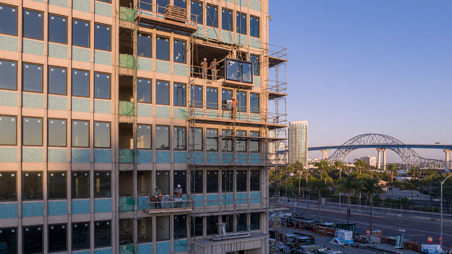 Downtown San Diego office tower undergoing conversion to residential use with construction scaffolding visible against a clear sky and Coronado Bridge in the distance