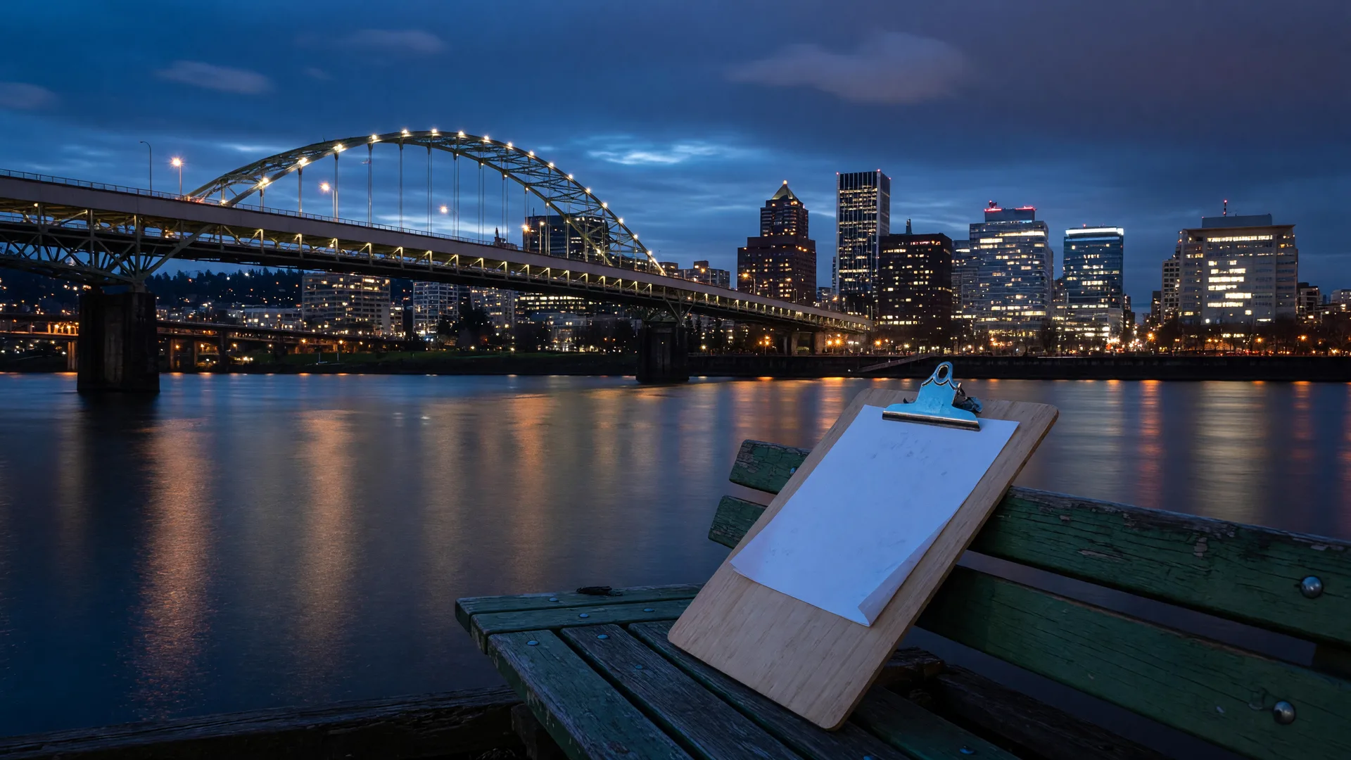The Portland skyline and Fremont Bridge at blue hour with a clipboard on a riverside bench