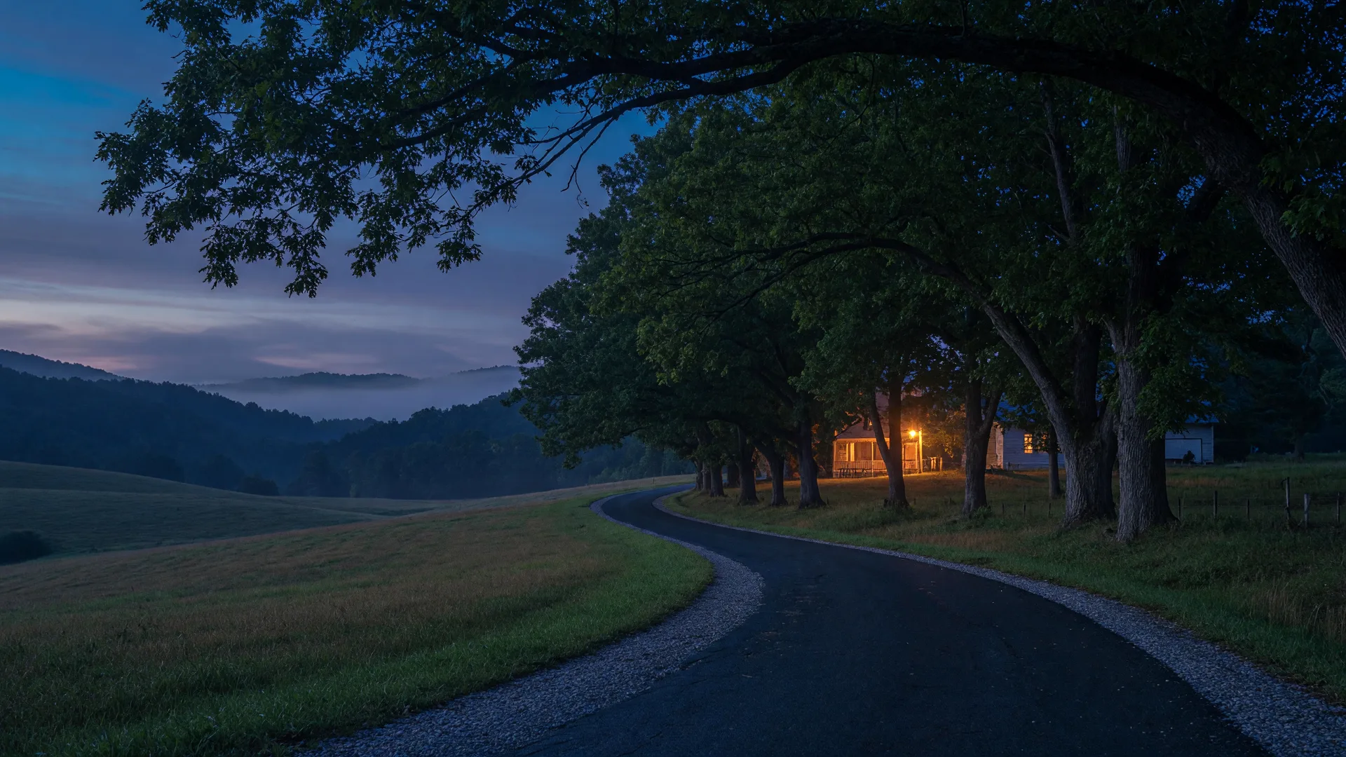 A quiet Kentucky country lane at blue hour with a warm farmhouse light through the trees