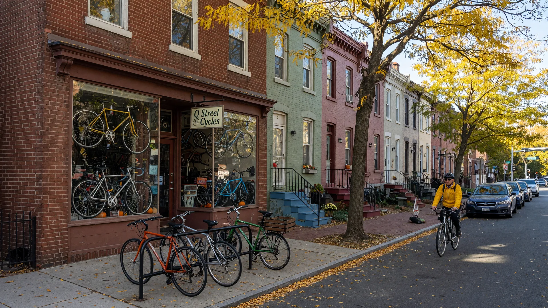 Classic two-story brick bike shop storefront on Q Street NW in the Logan Circle neighborhood of Washington DC on a bright late-October morning, display windows full of bicycles, a bike rack on the sidewalk, Victorian rowhouses behind, and a cyclist riding past under gold autumn trees